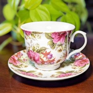 May include: A white teacup and saucer with a floral pattern featuring pink roses, green leaves, and purple berries. The cup has a handle and the saucer has a scalloped edge.