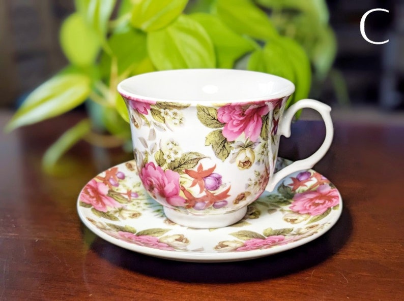 May include: A white teacup and saucer with a floral pattern featuring pink roses, green leaves, and purple berries. The cup has a handle and the saucer has a scalloped edge.