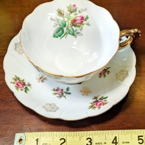 May include: A white teacup and saucer set with a gold trim and floral design. The cup features a pink rose with green leaves and the saucer has a scattered floral pattern with snowflakes.