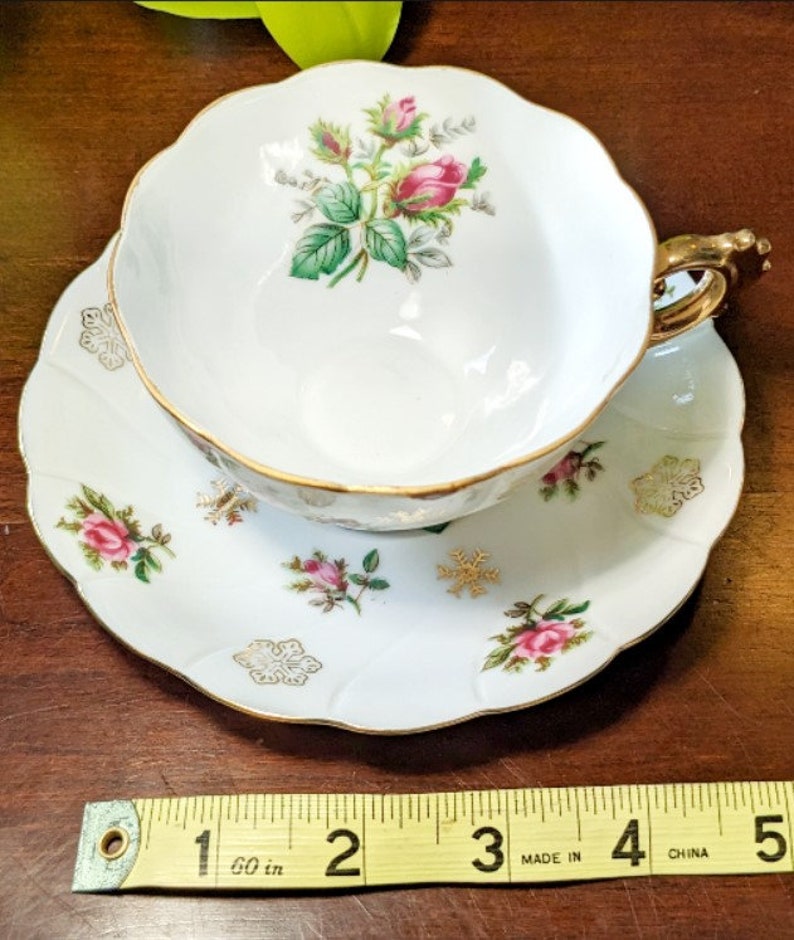 May include: A white teacup and saucer set with a gold trim and floral design. The cup features a pink rose with green leaves and the saucer has a scattered floral pattern with snowflakes.