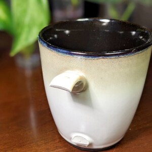 Può includere: Una tazza in ceramica bianca con un bordo nero e una glassa marrone chiaro. La tazza ha un design unico con un piccolo beccuccio laterale.
