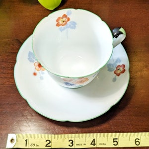 May include: A white teacup and saucer set with a green rim and floral design. The flowers are orange, red, and blue. The saucer has a scalloped edge.