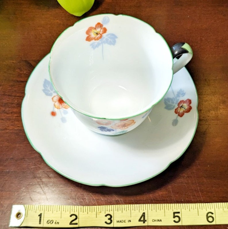 May include: A white teacup and saucer set with a green rim and floral design. The flowers are orange, red, and blue. The saucer has a scalloped edge.