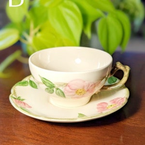 May include: A white teacup and saucer with a floral design featuring pink roses and green leaves. The cup has a handle shaped like a branch.