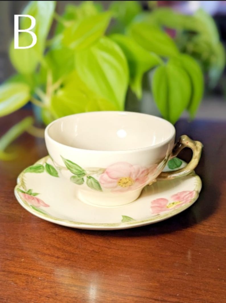 May include: A white teacup and saucer with a floral design featuring pink roses and green leaves. The cup has a handle shaped like a branch.