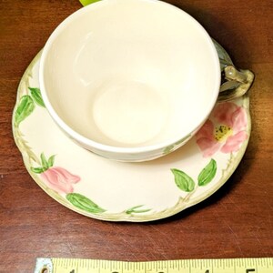 May include: A vintage teacup and saucer set with a floral design. The saucer is cream colored with a green vine and pink flower pattern. The teacup is cream colored with a green handle.
