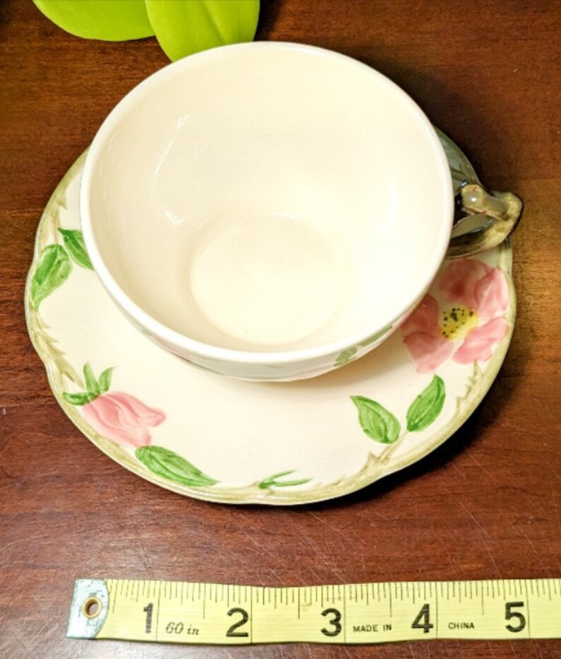 May include: A vintage teacup and saucer set with a floral design. The saucer is cream colored with a green vine and pink flower pattern. The teacup is cream colored with a green handle.