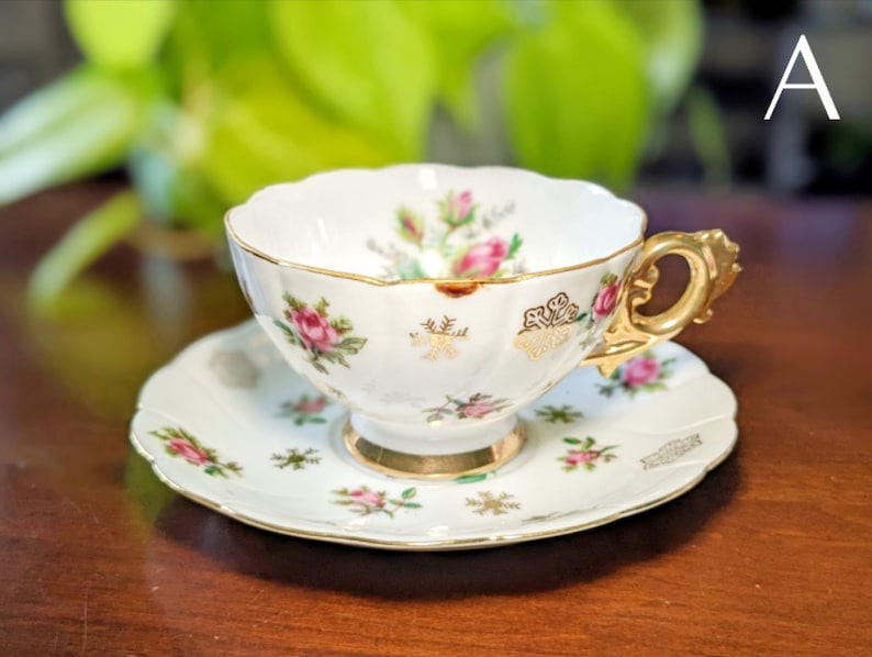 May include: A white teacup and saucer with a floral pattern and gold trim. The cup has a scalloped edge and a gold handle. The saucer has a matching floral pattern and gold trim.