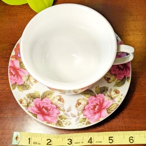 May include: A white teacup and saucer with a floral pattern. The saucer has pink roses and green leaves on a white background. The teacup is white with a gold rim. The saucer is 5 inches in diameter.