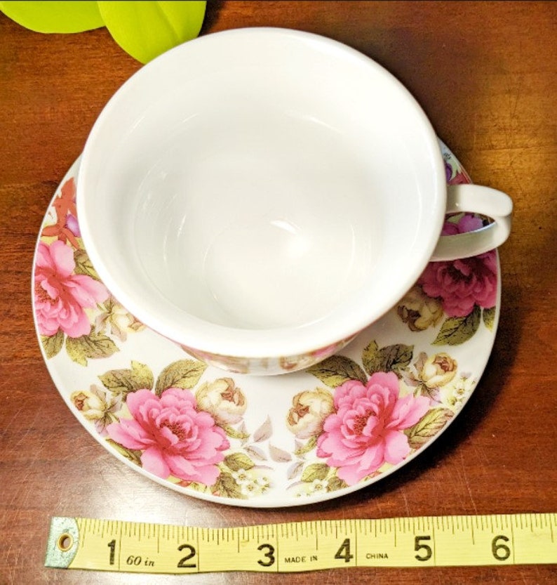 May include: A white teacup and saucer with a floral pattern. The saucer has pink roses and green leaves on a white background. The teacup is white with a gold rim. The saucer is 5 inches in diameter.