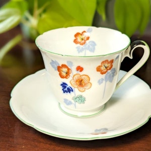 Può includere: Un set da tè bianco con tazza e piattino con un motivo floreale che presenta fiori arancioni, rossi, blu e verdi. La tazza ha un bordo verde e un manico. Il piattino ha un bordo ondulato.