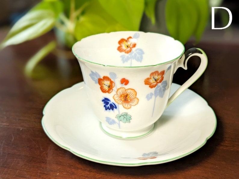 May include: A white teacup and saucer set with a floral design featuring orange, red, blue, and green flowers. The cup has a green rim and a handle. The saucer has a scalloped edge.