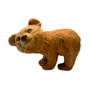 Puede incluir: Figura de oso de cerámica marrón con una expresión sonriente. El oso tiene una apariencia de pelaje texturizado, ojos pequeños y negros, y está posado en una postura juguetona. La figura es un objeto decorativo.
