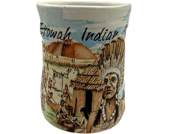 Może przedstawiać: Ceramiczny kubek ze szczegółową ilustracją osady rdzennych mieszkańców. Kubek przedstawia wizerunek indiańskiego wodza, domostw i napis "Etowah Indian Mounds" na tle błękitnego nieba. Kubek ma lekko nieregularny kształt.