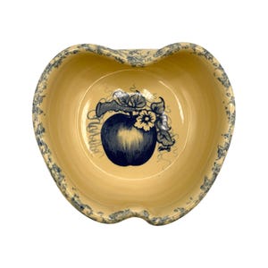 Può includere: Una ciotola in ceramica a forma di mela con interno giallo chiaro e un motivo floreale blu e bianco sul bordo. Il centro presenta un'illustrazione di mela blu con foglie e il nome "Sandy" in corsivo.