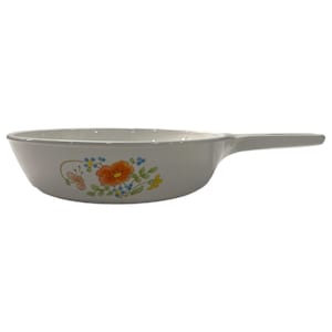 May include: White ceramic skillet with a long handle. The skillet features a floral design with orange, blue, and yellow flowers, and a butterfly. The skillet is designed for cooking and serving food.