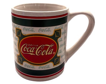 Op de afbeelding: Een witte keramische mok met een gebogen handvat. De mok heeft een rode ovaal met de woorden "Drink Coca-Cola" in witte letters. De mok heeft een zwarte en rode band rond de boven- en onderkant, met het Coca-Cola-logo herhaald.