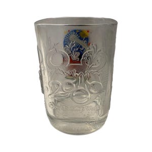 May include: Clear glass tumbler with embossed Disney World 2000 Celebration design. Features a colorful emblem with Mickey Mouse ears and fireworks. The glass has a square shape and is suitable for cold beverages.