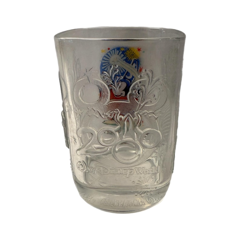 May include: Clear glass tumbler with embossed Disney World 2000 Celebration design. Features a colorful emblem with Mickey Mouse ears and fireworks. The glass has a square shape and is suitable for cold beverages.