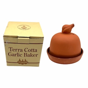 May include: A terracotta garlic baker with a dome-shaped lid featuring a garlic bulb-shaped handle, accompanied by its original beige cardboard box printed with the text Terra Cotta Garlic Baker and a graphic illustration of garlic bulbs.