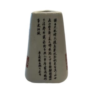 May include: A white ceramic vase with a slightly tapered shape. The vase features black calligraphy and red stamps. The calligraphy appears to be Asian characters. The vase is isolated on a white background.