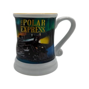 May include: A ceramic mug with a white handle and base, featuring a design from "The Polar Express." The mug's body displays a train illustration with the title in gold lettering against a dark, colorful background.