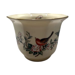 Peut inclure: Vase en céramique crème avec un bord évasé et une bordure dorée. Le vase présente une illustration détaillée d'un oiseau rouge perché parmi des fleurs roses et des feuilles vertes. Le motif se répète.