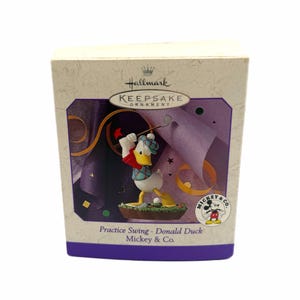 May include: Hallmark Keepsake ornament featuring Donald Duck in a golf outfit, mid-swing. The box has a purple and gold design with the text "Practice Swing - Donald Duck Mickey & Co."