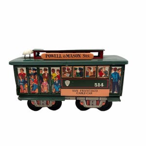 May include: A vintage green San Francisco cable car toy with the text "Powell & Mason Sts." and "San Francisco Cable Car." The toy features illustrations of passengers and the number 514.