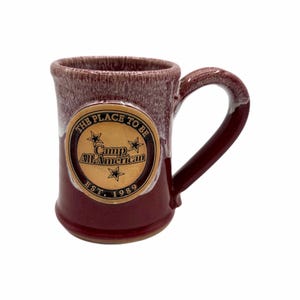 Puede incluir: Una taza de cerámica granate con asa. La taza presenta un emblema dorado con el texto "THE PLACE TO BE" y "Camp All-American EST. 1989". La taza tiene un acabado moteado y texturizado.