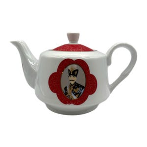 May include: White ceramic teapot with a red and gold design on the lid and a red quatrefoil frame on the body. The frame contains a portrait of a man in a military uniform. The teapot has a curved spout and handle.