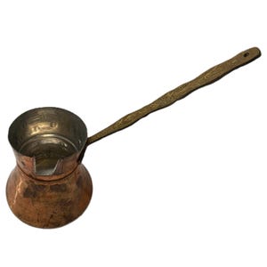 Peut inclure: Une cafetière en cuivre avec une longue poignée en laiton gravée. La cafetière a une base arrondie et un haut évasé.