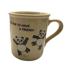 Peut inclure: Mug en céramique crème avec une anse incurvée. Le mug présente deux pandas de dessin animé sur des patins à roulettes et le texte "It's fun to have a friend !" en lettres noires. Le mug a un bord légèrement évasé et une finition lisse et brillante.