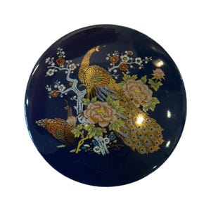 Puede incluir: Un plato decorativo redondo de color azul marino con un diseño detallado de pavo real y flores. El pavo real está representado con plumas doradas y marrones, posado en una rama florecida con flores color melocotón.