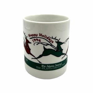 May include: A white ceramic mug with a black handle and the text "But First, Coffee" in a bold, black font. The mug is set against a blurred background, suggesting a cozy setting.