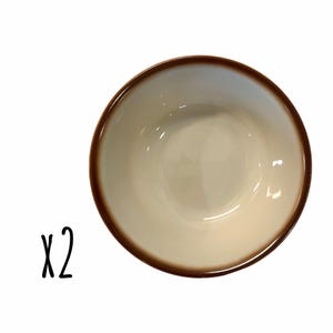 May include: Two off-white ceramic bowls with a brown rim. The bowls have a glossy finish and are set against a white background. The text "x2" is written in black script next to the bowls.