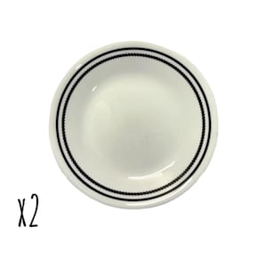 May include: Two small, white ceramic plates with a black dotted border. The plates are round and have a slightly concave center. The text "x2" is written in black script in the lower left corner.
