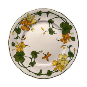 Può includere: Piatto in ceramica bianca con bordo smerlato, decorato con un motivo floreale. Il motivo presenta fiori gialli e arancioni, foglie verdi e steli. Il bordo è delineato da un bordo verde.