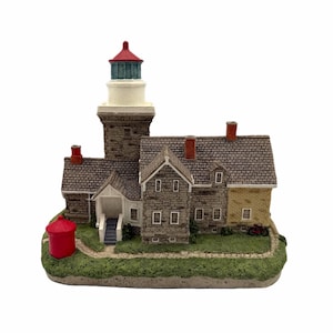 Könnte beinhalten: Ein detailliertes Miniatur-Leuchtturmmodell mit einem rot gekrönten Turm, einem weißen Licht und einem steintexturierten Sockel. Das Modell umfasst ein Haus mit einer grauen Steinfassade, einen roten Wasserturm und einen kleinen Pfad.