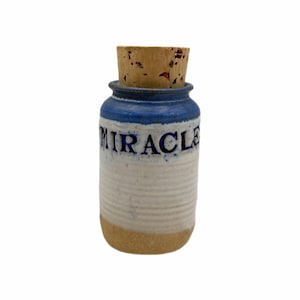 Peut inclure: Petit pot cylindrique en céramique avec bouchon en liège. Le pot est blanc avec une bande bleue en haut et une base beige. Le mot "MIRACLE" est imprimé en bleu sur le devant.