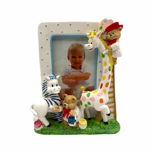 May include: A decorative baby photo frame featuring a smiling baby. The frame is adorned with a zebra, a teddy bear, and a giraffe. The giraffe has colorful spots, and the teddy bear is holding a paint bucket. The frame has a blue border.