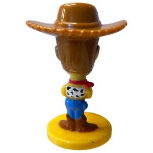 Może przedstawiać: Plastikowa figurka Woody'ego z Toy Story. Woody ma na sobie sw&oacute;j charakterystyczny brązowy kowbojski kapelusz, niebieskie dżinsy i ż&oacute;łtą koszulę z czerwoną bandaną. Stoi na ż&oacute;łtej podstawie.