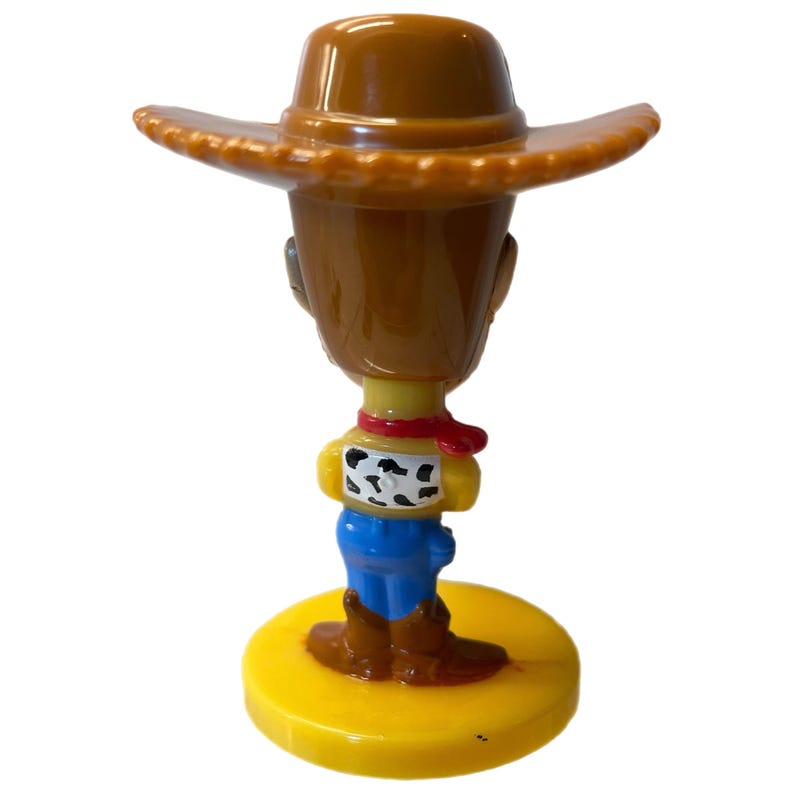 Może przedstawiać: Plastikowa figurka Woody'ego z Toy Story. Woody ma na sobie sw&oacute;j charakterystyczny brązowy kowbojski kapelusz, niebieskie dżinsy i ż&oacute;łtą koszulę z czerwoną bandaną. Stoi na ż&oacute;łtej podstawie.