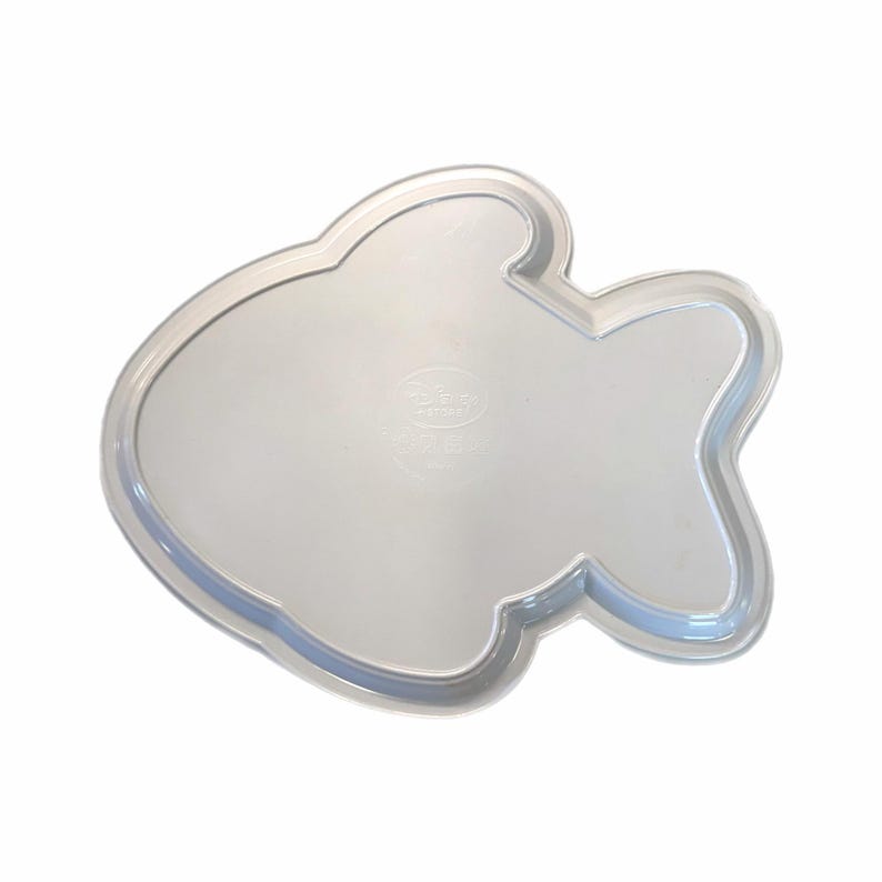 May include: A clear, fish-shaped baking pan. The pan has a smooth, reflective surface and a slightly raised edge. The pan is made of a clear material, and the design is visible from the top.