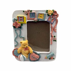 May include: A white rectangular picture frame with the word "BABY" spelled out in colorful blocks. The frame is decorated with a yellow teddy bear, pink and blue ribbons, and small flowers. A heart-shaped pillow with the word "LOVE" is also included.