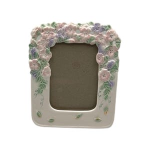 Può includere: Una cornice per foto in ceramica bianca con apertura rettangolare. La cornice è decorata con un motivo floreale rosa, lilla e verde. La cornice ha una finitura lucida ed è adatta per esporre una fotografia.