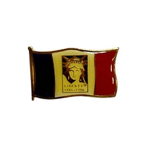 Op de afbeelding: Een rechthoekige speld met een ontwerp van de Franse vlag met een gouden rand. Het middenpaneel toont het Vrijheidsbeeld met de tekst "LIBERTY 1886-1986". De speld heeft een gouden rand en staat tegen een witte achtergrond.