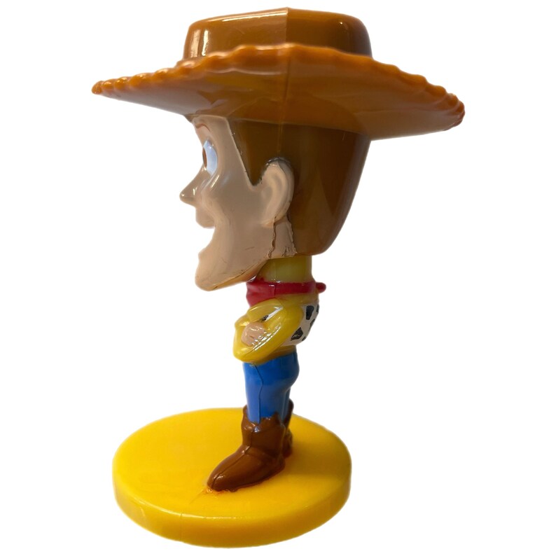Może przedstawiać: Plastikowa figurka Woody'ego z Toy Story. Woody ma na sobie sw&oacute;j charakterystyczny brązowy kowbojski kapelusz, ż&oacute;łtą kamizelkę, czerwoną chustkę i niebieskie dżinsy. Stoi na ż&oacute;łtej podstawie.