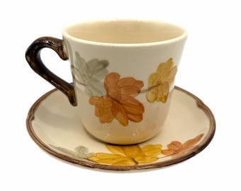 May include: Cream-coloured teacup and saucer set with a brown handle and rim. The cup and saucer are decorated with autumn-coloured leaf designs in shades of orange, yellow, and green. The cup sits on a matching saucer.