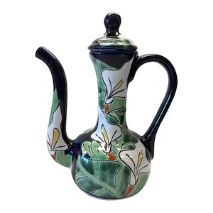Peut inclure: Pichet en céramique avec un corps long et élancé et un bec verseur incurvé. Le pichet est décoré de callas blanches, de feuilles vertes et d'accents jaunes et oranges sur un fond bleu foncé. Il a un couvercle assorti et une poignée incurvée.
